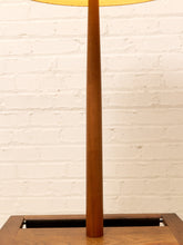 Load image into Gallery viewer, A close-up of a mid-century modern floor lamp with a tapered wooden pole and a warm yellow fabric shade, set against a white painted brick wall.
