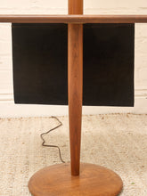 Load image into Gallery viewer, A mid-century modern wooden floor lamp with a warm brown wood finish, featuring a round base, a central pole, and a black fabric shade, shown on a textured beige rug against a white brick wall.
