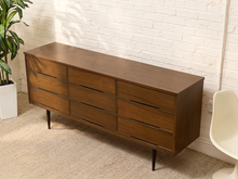 Load image into Gallery viewer, Mid-century modern wooden dresser with nine drawers, shown in a styled room setting with a white brick wall, beige carpet, and a potted plant.

