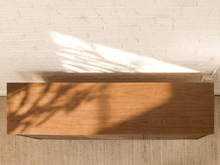 Load image into Gallery viewer, A rectangular wooden floating shelf with a warm medium-brown finish, mounted on a white brick wall above a textured beige rug. Natural light and leaf shadows are cast across the shelf and wall.
