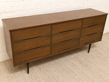 Load image into Gallery viewer, A mid-century modern six-drawer wooden dresser with a warm brown walnut finish and slim metal pull handles, shown on a textured beige carpet against a white brick wall.

