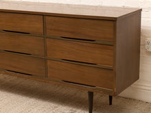 Load image into Gallery viewer, Mid-century modern wooden dresser with six drawers, dark metal handles, and tapered legs, shown in a room setting against a white brick wall and carpet.
