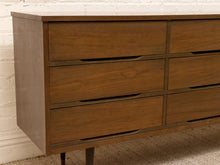 Load image into Gallery viewer, Mid-century modern wooden dresser with six drawers, dark metal handles, and tapered legs, shown against a white brick wall and carpeted floor.
