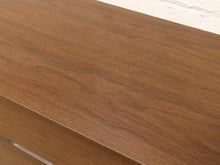 Load image into Gallery viewer, Close-up of a medium brown wood-grain surface, likely a dresser or cabinet front, with a white painted brick wall visible in the background.
