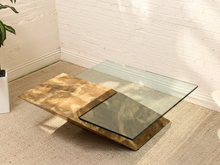 Load image into Gallery viewer, A modern square coffee table with a textured gold and bronze metal base, a dark smoked glass front panel, and a clear glass top, set on a beige carpet against a white brick wall.
