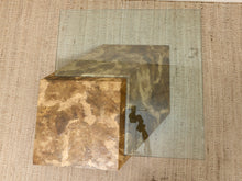 Load image into Gallery viewer, A modern square coffee table with a clear glass top and two textured metal side panels in a mottled bronze and gold finish, shown on a woven beige rug.
