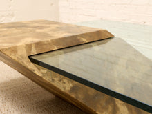 Load image into Gallery viewer, A close-up of a modern coffee table with a thick, wavy organic-shaped metallic gold and brown base, a triangular glass panel, and a rectangular stone slab lid, set against a white brick wall background.
