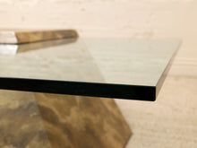 Load image into Gallery viewer, Close-up of a modern glass tabletop with a thick black edge and a reflective gold-toned base, set against a light beige wall and floor.
