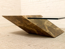 Load image into Gallery viewer, A modern sculptural coffee table with a geometric wedge base in a mottled bronze and brown finish, supporting a clear glass top, set against a white brick wall and beige carpet.
