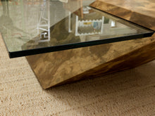 Load image into Gallery viewer, A close-up of a modern coffee table with a thick, angular wedge-shaped base made of polished brown wood grain and a clear glass top, resting on a beige woven rug.
