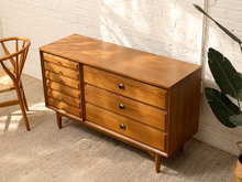 Load image into Gallery viewer, A mid-century modern wooden sideboard with six drawers, shown in a styled room setting with a white brick wall, beige carpet, a wooden chair, and a potted plant.
