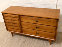 Load image into Gallery viewer, Mid-century modern wooden dresser with a warm medium-brown finish, featuring a column of five small drawers on the left, three larger drawers on the right, and round brass pulls, set against a white brick wall and textured rug.

