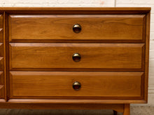 Load image into Gallery viewer, A mid-century modern wooden dresser with three drawers, featuring a warm medium-brown wood finish and round dark metal pulls, set against a white brick wall background.
