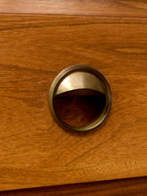Load image into Gallery viewer, A close-up of a round brass drawer pull handle mounted on a warm brown wooden cabinet or drawer front.
