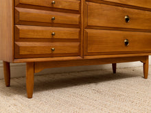Load image into Gallery viewer, Mid-century modern wooden dresser with a warm honey finish, featuring multiple drawers with round brass pulls and tapered legs, shown on a beige carpeted floor.
