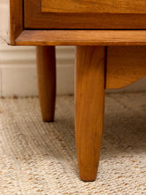 Load image into Gallery viewer, Close-up of a mid-century modern wooden side table or cabinet leg, showing the warm medium-brown wood grain and tapered leg, resting on a woven jute rug.
