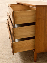 Load image into Gallery viewer, A mid-century modern wooden chest of drawers with three open drawers, showcasing a warm honey-toned wood grain finish and brass drawer pulls, set against a neutral carpet and white brick wall background.
