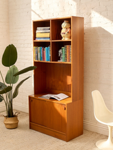 Load image into Gallery viewer, A mid-century modern wooden bookcase with a warm honey finish, styled with books and a ceramic figurine against a white brick wall. A potted plant and a white molded chair are visible in the room.
