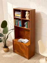 Load image into Gallery viewer, A mid-century modern wooden bookcase with a warm cherry finish, styled with books, a ceramic figurine, and an open magazine. It is set against a white brick wall with a potted plant on the left and a white chair on the right.
