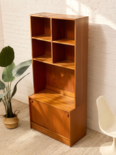 Load image into Gallery viewer, A mid-century modern wooden bookcase with six open shelves, a lower cabinet door, and a slanted top, shown in a styled room corner with a white brick wall, potted plant, and chair.
