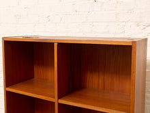 Load image into Gallery viewer, A close-up of a wooden bookshelf with a warm medium-brown finish, showing two open cubbies and a top shelf, set against a white painted brick wall.
