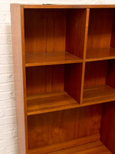 Load image into Gallery viewer, A close-up of a wooden bookshelf with a warm reddish-brown finish, showing multiple open cubby compartments, photographed against a white painted brick wall.
