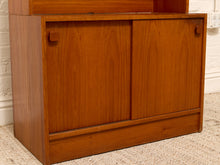 Load image into Gallery viewer, A mid-century modern wooden cabinet with two sliding doors, warm medium-brown teak wood grain, and round wooden knobs, set against a white brick wall and beige carpet.
