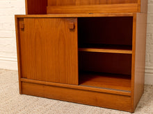 Load image into Gallery viewer, A mid-century modern wooden cabinet with a warm honey-toned finish, featuring a sliding door and open shelving, set against a white brick wall and beige carpet.
