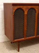Load image into Gallery viewer, A mid-century modern wooden credenza or sideboard cabinet with a warm medium-brown wood finish, featuring two arched cane or wicker screen panels on the front and tapered legs, shown against a white brick wall and beige carpet.
