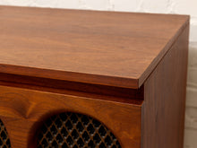 Load image into Gallery viewer, Close-up of a wooden cabinet or speaker with a warm medium-brown wood finish and a dark mesh grille, set against a white brick wall background.
