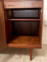 Load image into Gallery viewer, A mid-century modern wooden cabinet with an open door, showcasing its warm brown wood grain, interior shelves, and black metal wire supports, set against a white brick wall and textured carpet.
