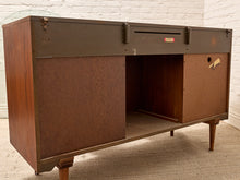 Load image into Gallery viewer, A vintage mid-century modern wooden writing desk or console table with a dark metal top and reddish-brown wood sides and drawer fronts, shown in a room with a white brick wall and carpeted floor.
