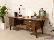 Load image into Gallery viewer, A mid-century modern wooden writing desk with a warm brown finish and silver drawer pulls, styled in a home office setting with a lamp, books, a plant, and a clock against a white brick wall.
