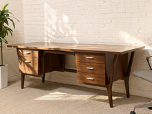 Load image into Gallery viewer, Mid-century modern wooden writing desk with a warm brown finish, featuring three drawers on the left, three drawers on the right, and angled legs, shown in a room setting with a white brick wall and beige carpet.
