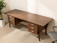 Load image into Gallery viewer, A mid-century modern wooden writing desk with a warm brown finish, featuring three drawers on each end with silver handles, set against a white brick wall and beige carpet.

