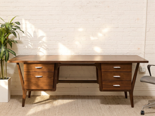 Load image into Gallery viewer, A mid-century modern wooden writing desk with a warm brown finish, featuring three drawers on each end and a central open space, set against a white brick wall with sunlight filtering through.
