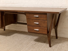 Load image into Gallery viewer, Mid-century modern wooden writing desk with three drawers, silver handles, and angled legs, set against a white brick wall and beige carpet.
