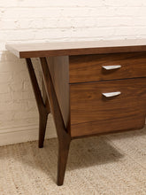 Load image into Gallery viewer, Mid-century modern wooden nightstand with two drawers, silver handles, and angled legs, positioned against a white brick wall on a textured beige rug.
