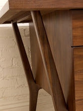 Load image into Gallery viewer, Close-up detail of a mid-century modern wooden desk, showing the rich grain of dark walnut wood on the drawer front, side panel, and legs, with a light brick wall in the background.
