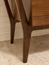 Load image into Gallery viewer, Close-up of a mid-century modern wooden sideboard or cabinet leg, showcasing a rich walnut finish, set against a light brick wall and textured beige rug.
