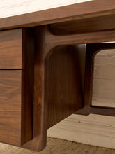 Load image into Gallery viewer, Close-up of a dark walnut wood desk showing the drawer front, apron, and curved leg support structure against a light brick wall background.
