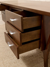 Load image into Gallery viewer, Close-up of a mid-century modern wooden desk with three open drawers showing silver handles, set on a textured beige carpet.
