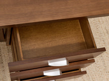 Load image into Gallery viewer, Close-up of a wooden desk drawer pulled open, showing a medium brown wood finish and white drawer pulls on a textured beige rug.
