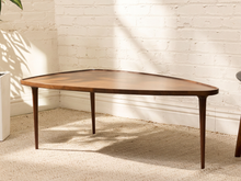 Load image into Gallery viewer, A mid-century modern wooden coffee table with a rounded triangular top and tapered legs, set against a white brick wall on a beige carpet.
