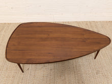 Load image into Gallery viewer, A mid-century modern teardrop-shaped wooden coffee table with a rich walnut finish, shown on a woven jute rug against a white brick wall.
