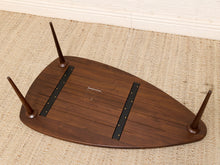 Load image into Gallery viewer, A dark brown wooden table base or stool bottom with four tapered legs and two black metal strip brackets, photographed upside down on a beige woven rug.

