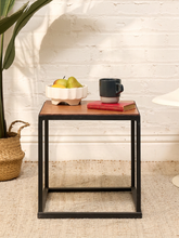 Load image into Gallery viewer, A square side table with a brown wood top and a black metal frame, styled with a bowl of pears, a mug, and a book on a woven rug against a white brick wall.
