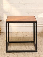 Load image into Gallery viewer, A square side table with a warm brown wood-grain top and a black metal frame, set on a beige textured rug against a white brick wall.
