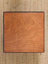 Load image into Gallery viewer, A square wooden board or tile with a warm medium-brown wood grain finish and a thin dark border, placed on a woven beige textured surface.
