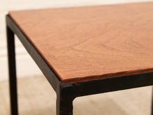 Load image into Gallery viewer, Close-up of a square wooden table top with a warm medium-brown wood grain finish and a black metal frame leg, shown against a light neutral background.
